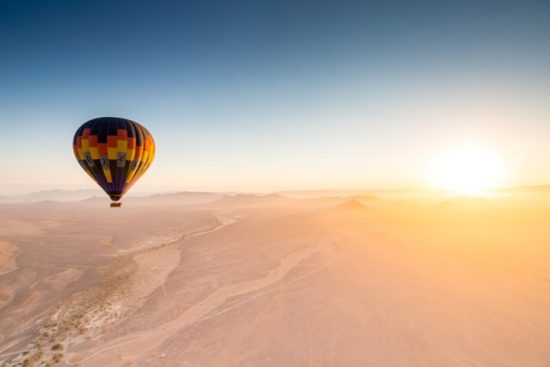 Ein Heißluftballon fliegt über die Wüste gen aufgehender Sonne